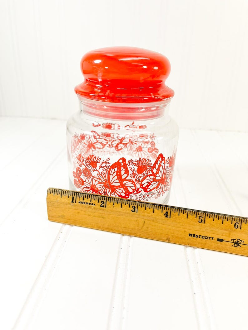 May include: A clear glass jar with a red lid and a red butterfly and floral design. The jar is approximately 4 inches tall. A wooden ruler is placed next to the jar.