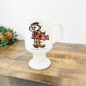 May include: White pedestal mug with a cartoon bear riding a scooter. The bear wears a crown and red and white striped pants. The mug has a handle and the text "RINGO" and "CAROWINDS CORPORATION" printed on it. The mug is on a wooden surface.