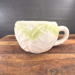 May include: A white ceramic mug shaped like a head of cabbage with green leaves. The mug has a handle on the side.