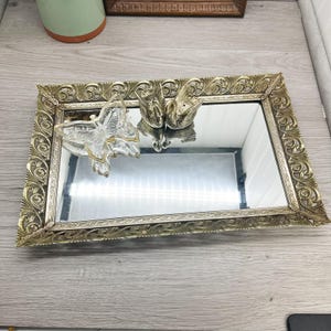 Könnte beinhalten: Ein verziertes Tablett in Goldtönen mit einer Spiegel Oberfläche. Das Tablett zeigt einen Glas-Schmetterling und zwei goldfarbene Vögel.
