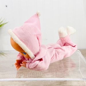 Puede incluir: Una muñeca bebé vintage con un conjunto de forro polar rosa y un gorro a juego con ribete blanco. La muñeca tiene una cara color melocotón, ojos azules y manos y pies pequeños. La muñeca está tumbada boca abajo.