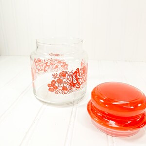 May include: A clear glass jar with a red lid, decorated with red floral and butterfly designs. The jar is open, with the lid placed to the side. The design is a vintage style.