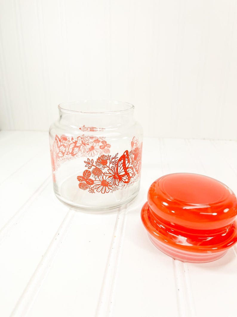 May include: A clear glass jar with a red lid, decorated with red floral and butterfly designs. The jar is open, with the lid placed to the side. The design is a vintage style.