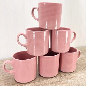 May include: Six pink ceramic mugs stacked in a pyramid shape. Each mug has a handle and a ribbed texture. The mugs are a solid, soft pink color and appear to be empty.