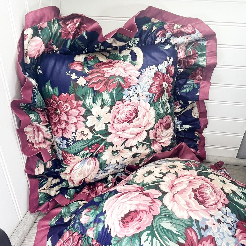 May include: Two decorative cushions with a floral pattern. The cushions feature a navy blue background with pink, red, and white flowers. The edges are trimmed with a ruffled, dark pink satin.