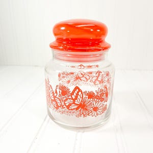 May include: A clear glass jar with a bright orange lid and a decorative floral and butterfly design in orange. The jar is cylindrical with a rounded top and a screw-on lid. The design wraps around the jar.