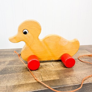 May include: A wooden pull-along toy duck with a light brown body, black eye, and red wheels. The duck has a long orange string attached for pulling. The toy is made of wood and has a classic, vintage design.