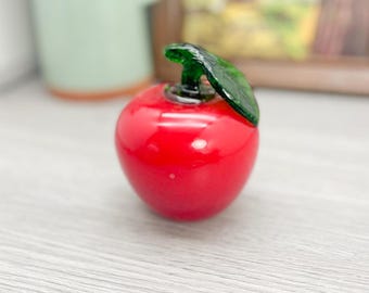 May include: A red glass apple with a green glass stem and leaf. The apple is sitting on a light gray surface.