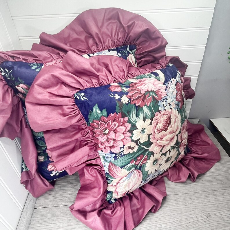 May include: Cushions with a floral design on a navy blue base. The cushions have a ruffled, mauve-coloured border. The floral pattern includes pink, white, and green flowers. These cushions are for home decor.