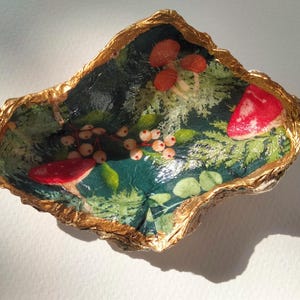 May include: An oyster shell dish with a gold-painted edge. The interior is decorated with a forest scene, including red mushrooms, berries, and green leaves. Ideal for holding small items.