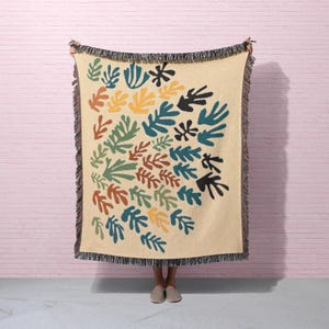 May include: A beige throw blanket with a colorful leaf pattern in shades of green, blue, yellow, orange, and black. The blanket has a fringed edge and is held up against a pink brick wall.