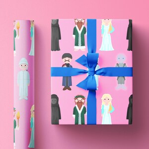 Puede incluir: Papel de regalo rosa y un regalo envuelto con una cinta azul. El papel presenta ilustraciones repetidas de personajes con varios atuendos. Un rollo de papel a juego está a la izquierda. El regalo está envuelto con una cinta azul.