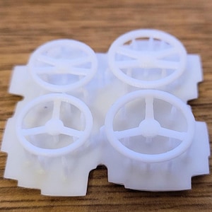 May include: Four small, white plastic steering wheel miniatures. Each wheel has a circular rim with spokes connecting to a central hub. The wheels are attached to a white, irregularly shaped base.