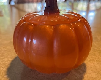 May include: A round, orange pumpkin-shaped container with a smooth, glossy finish and a dark brown stem. The object features vertical ridges and a lid that fits into the top, creating a segmented appearance with a subtle shimmering effect.
