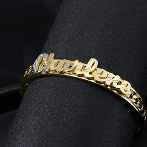 Op de afbeelding: Een goudkleurige armband met de naam "Charlotte" in een cursief lettertype. De armband heeft een schakelontwerp en wordt op een zwart oppervlak weergegeven. De naam heeft een contrasterende kleur.
