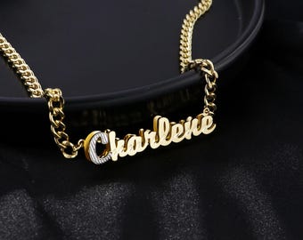 Può includere: Una collana color oro con il nome "Charlene" in corsivo. La collana ha un design a catena a maglia. Il nome è in uno stile a strati con un accento color argento.