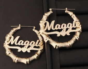 Puede incluir: Pendientes de aro dorados con el nombre "Maggie" escrito en letra cursiva en cada pendiente. Los pendientes están hechos de un material similar al bambú.