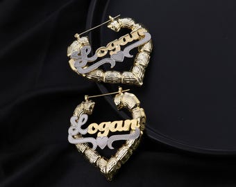 May include: A pair of gold-tone heart-shaped hoop earrings with the name "Logan" in a cursive font. The earrings have a two-tone design, with silver-tone accents. The earrings are displayed on a black surface.