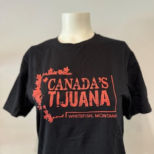 May include: Black t-shirt with the text "CANADA'S TIJUANA" in red, along with "WHITEFISH, MONTANA" below. The design includes red maple leaf accents, creating a bold graphic.