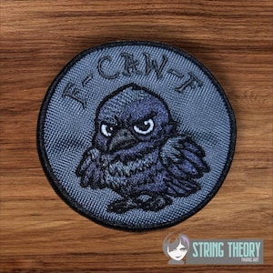 May include: A circular patch featuring a cartoon raven with an angry expression. The raven is dark blue with white eyes and is centered on a gray background. The text "F-CAW-F" is arched above the raven. The patch has a black border.