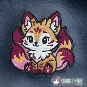 May include: Embroidered patch featuring a cartoon fox with multiple tails. The fox has a white face, orange and brown fur, and pink and burgundy accents. The patch is outlined in black and includes the text "STRING THEORY FABRIC ART".