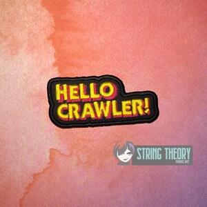 May include: A rectangular embroidered patch with a black border and black background. The text HELLO CRAWLER! is stitched in bright yellow with a pink shadow effect, creating a bold, layered appearance across the center of the fabric patch.
