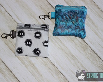 May include: Two small zippered pouches with keychains. One pouch is white with a black and white pattern of cartoon eyes. The other pouch is blue with a swirling marbled pattern. The text "STRING THEORY FABRIC ART" is visible in the bottom right corner of the image.