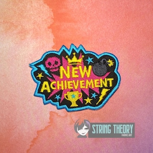 May include: An embroidered patch with a black background and a bright blue border. The text NEW ACHIEVEMENT is in yellow. The design features a pink skull, a yellow crown, a yellow trophy, a black bomb, and yellow and blue lightning bolts and stars.