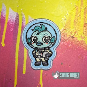 May include: An embroidered patch featuring a light blue aquatic creature with large eyes wearing a space helmet and a beige spacesuit. The character has teal hair and is holding a small gray object. The patch has a light blue border.