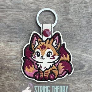 May include: Embroidered fox keychain with a white background, featuring a cute fox design in shades of orange, brown, and burgundy. The keychain has a metal ring and a white leather strap with a burgundy button. The text "STRING THEORY FABRIC ART" is visible.