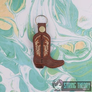 May include: Brown leather boot keychain with a gold key ring. The boot has a brown and orange embroidered design.
