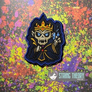 May include: An embroidered patch featuring a skeletal figure wearing a gold crown with red accents, dressed in a dark blue and gold robe. The figure has glowing blue eyes and holds a small blue flame in one hand, all outlined in dark blue.