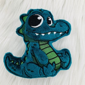 May include: A teal plush alligator toy with a green belly and accents. The alligator has large white eyes, a stitched mouth with white teeth, and a green outline. The toy is made of soft, textured fabric.