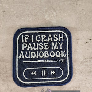 以下が含まれることがあります： 「IF I CRASH PAUSE MY AUDIOBOOK」と書かれた白い文字が刺繍された、濃いネイビーブルーのパッチ。パッチには再生/一時停止ボタンと巻き戻し/早送りボタンが付いています。右下隅には「STRING THEORY FABRIC ART」という文字があります。