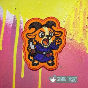 May include: An embroidered patch featuring a cartoon goat character with orange fur, black horns, and a white chest patch. The goat wears a dark blue robe with a brown belt and holds a small brown bottle. The patch has a thick orange border.
