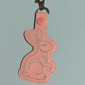 May include: A pink leather keychain with a silver embroidered bunny design. The keychain has a silver metal ring and a pink button.
