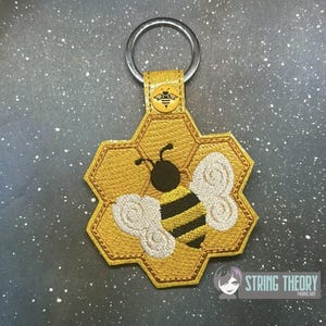 May include: A gold-colored, honeycomb-shaped keychain with a bee design. The bee has black and yellow stripes and white wings. The keychain has a metal ring and a small bee emblem on the strap.