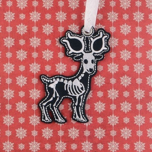 以下が含まれることがあります： 白黒のトナカイの骨格のクリスマスオーナメント。白いリボンが付いており、吊り下げられます。トナカイは白い糸で縫われ、詳細な骨格のデザインが施されています。背景は白い雪の結晶の赤いパターンです。オーナメントはファブリックアートです。