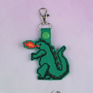 May include: Green pixelated dragon keychain with a red flame coming out of its mouth. The keychain has a silver metal clasp and a green button.