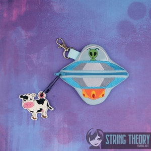 May include: A blue and gray UFO-shaped zippered pouch with a green alien inside. The pouch is attached to a black and silver chain with a cow charm.