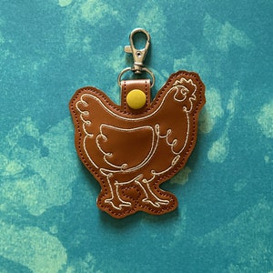 May include: Brown leather chicken keychain with a yellow button and a silver metal clasp. The chicken is outlined in white thread.