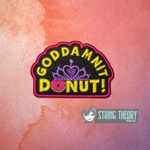 Peut inclure: Un écusson brodé avec un fond noir et une bordure rose. Le texte GODDAMNIT DONUT! est brodé en jaune. Un diadème violet est placé au-dessus d'un donut rose avec des vermicelles jaunes, remplaçant la lettre O du mot DONUT.