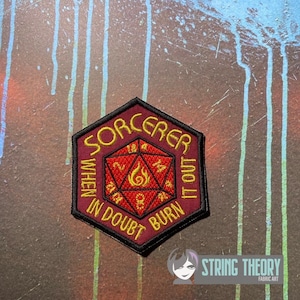 May include: A hexagonal embroidered patch with a dark red background and a black border. The center features a red twenty-sided die with yellow numbers and a flame icon. The yellow text reads SORCERER WHEN IN DOUBT BURN IT OUT.