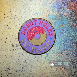 May include: A circular embroidered patch with a purple background and a dark brown border. The center features a tan donut with pink icing and blue sprinkles. The text reads DONUT HOLES EST. FLOOR 1 in gold and red stitched lettering.