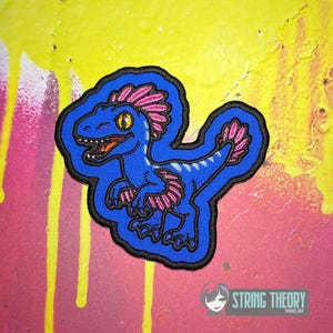May include: An embroidered patch featuring a blue dinosaur with pink feathers on its head, arms, and tail. The dinosaur has a yellow eye and an open mouth showing teeth. The patch has a thick black border and is set against a yellow and pink background with the text STRING THEORY FABRIC ART in the corner.