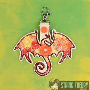 Puede incluir: Un llavero de dragón bordado en rojo y naranja sobre un fondo blanco con un anillo de llave plateado. El dragón está enroscado con las alas extendidas. El llavero está hecho por String Theory Fabric Art.