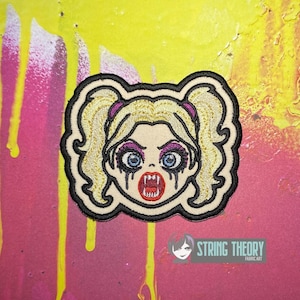 May include: An embroidered patch featuring a stylised character with blonde pigtails, blue eyes, purple eyeshadow, and sharp fangs. The design includes black outlines, a cream-coloured face, and a red mouth. The patch has a black border.