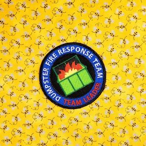 Op de afbeelding: Gele stof met een herhaald patroon van bijen. Een cirkelvormige patch met de tekst "DUMPSTER FIRE RESPONSE TEAM" omringt een groene vuilnisbak met vlammen en de woorden "TEAM LEADER".