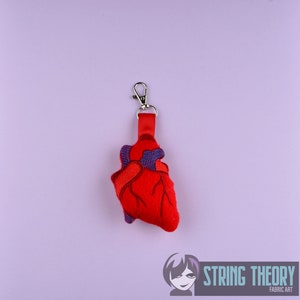 Könnte beinhalten: Schlüsselanhänger aus rotem Filz in Form eines anatomischen Herzens mit violetten Nähten und einem silbernen Metallverschluss. Der Schlüsselanhänger hängt an einem roten Band.