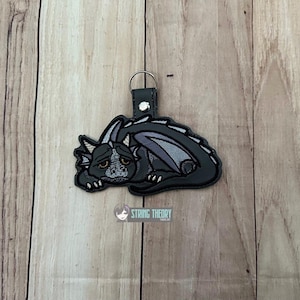 May include: A black dragon keychain with a sad expression. The dragon has gray wings and accents, and is lying down. The keychain has a black leather strap and a silver-colored ring. The String Theory logo is visible.